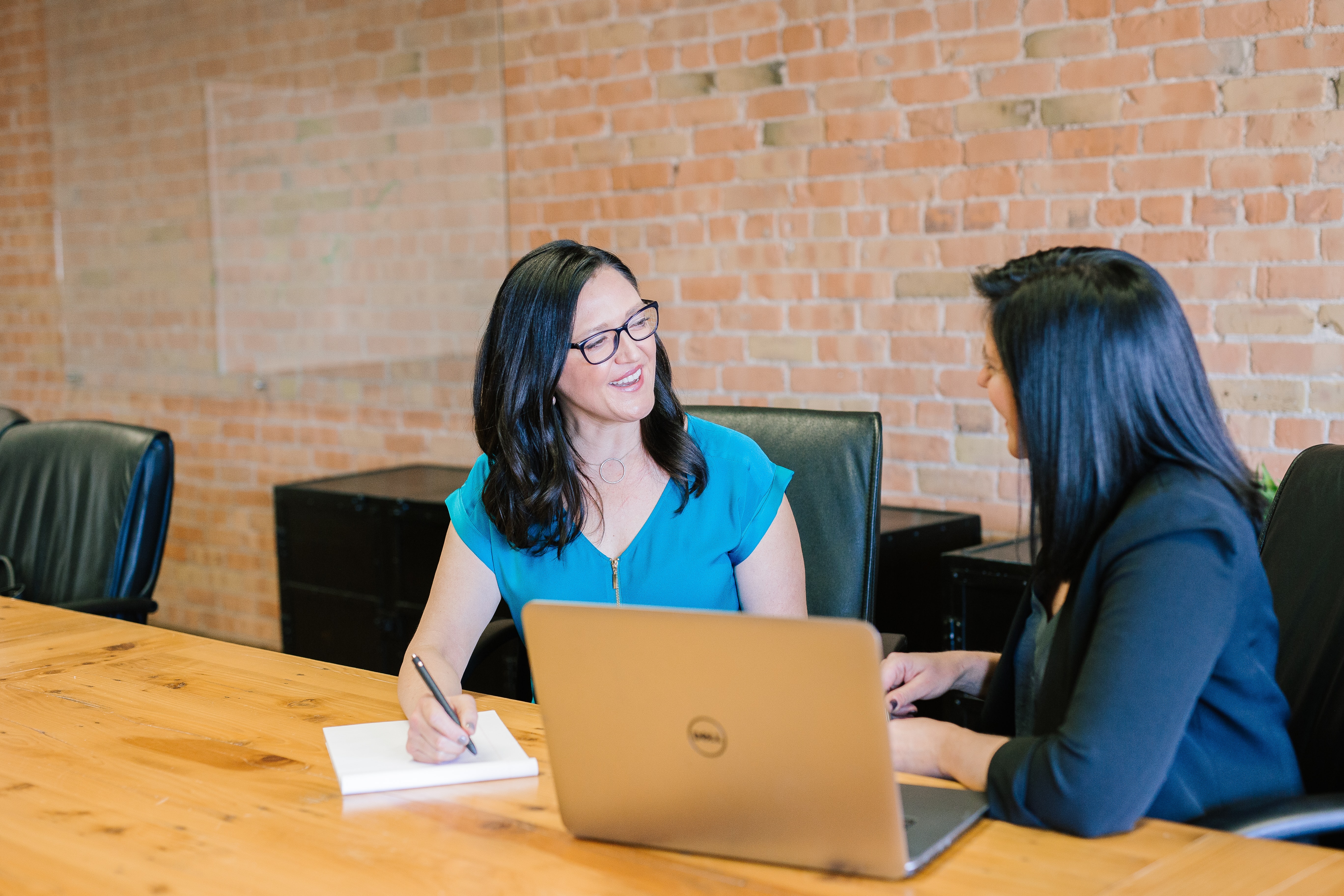 two women sat at table with laptop and notepad and pen working together smiling